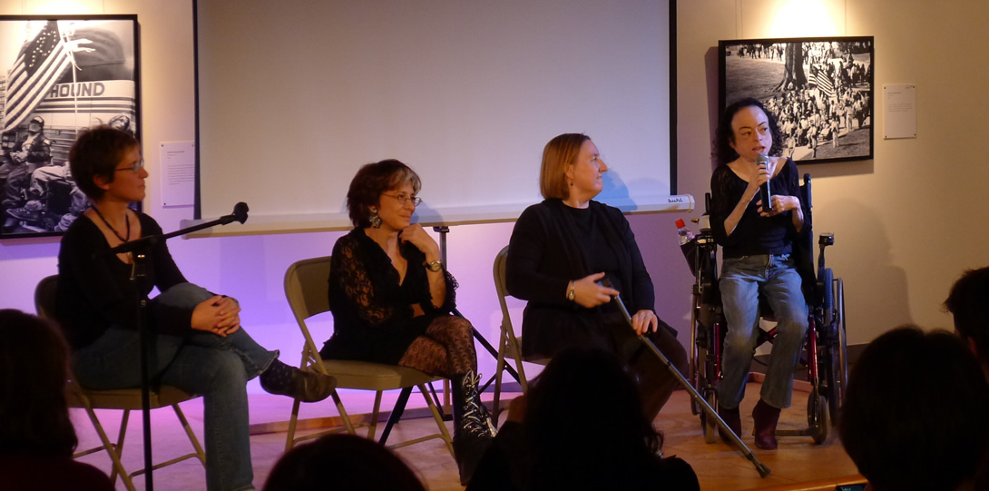 Disabled Artists and Activists Riva Lehrer, Carrie Sandahl, Liz Carr and another woman in active discussion. The women sit in a semi-circle. From left to right, a white woman with short-cropped brown hair and glasses sits in a chair with her legs crossed listening to the speaker. She wears jeans and a black long sleeve shirt. Next to her is a white woman with short, cropped grey and red hair and glasses. She is seated with her legs crossed also listening to the speaker. She wears a black lace patterned dress, patterned black tights and orthotic black boots that resemble combat boots, tied with silver sparkly laces. Next is a seated white woman with shoulder length blonde hair, dressed in black and holding a cane, listening to the woman to her right. The woman speaking is white and sits in her wheelchair holding a microphone in her hand. She has a small frame and curly shoulder length brown hair.