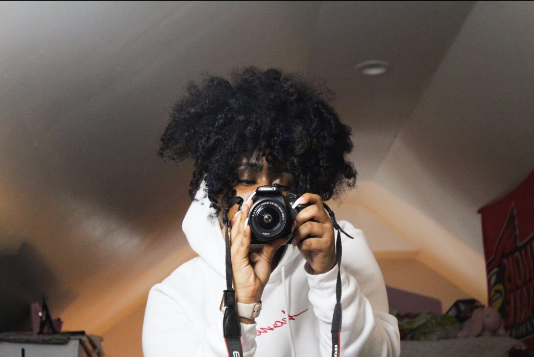A black woman taking a picture in the mirror with her black canon camera.she has on a white hoodie, and her hair is in a high ponytail.
