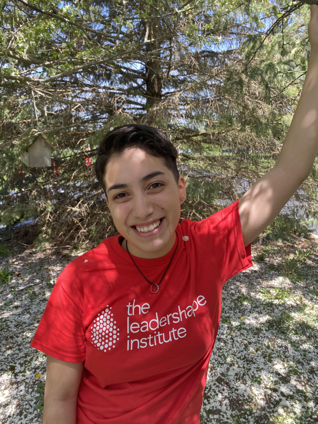 Febe is a Latinx person in a red short sleeve shirt smiling at the camera with one arm raised. Behind them, there is a large pine tree.