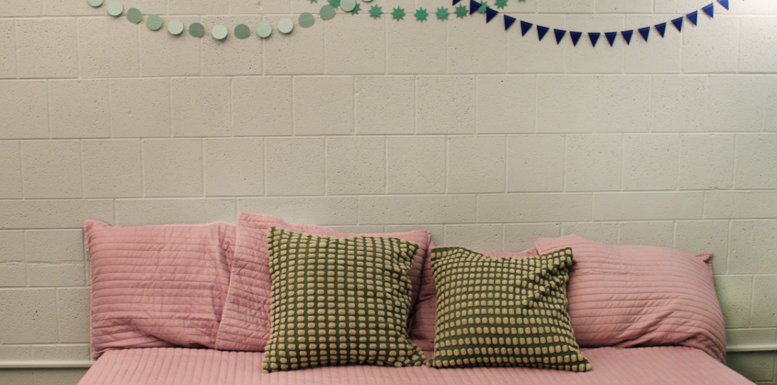 A light blue wall with three garlands hung on it towards the top of the picture. The first garland is light blue, the second is green, and the third is dark blue. There is a pink daybed in the center with pink and green pillows.