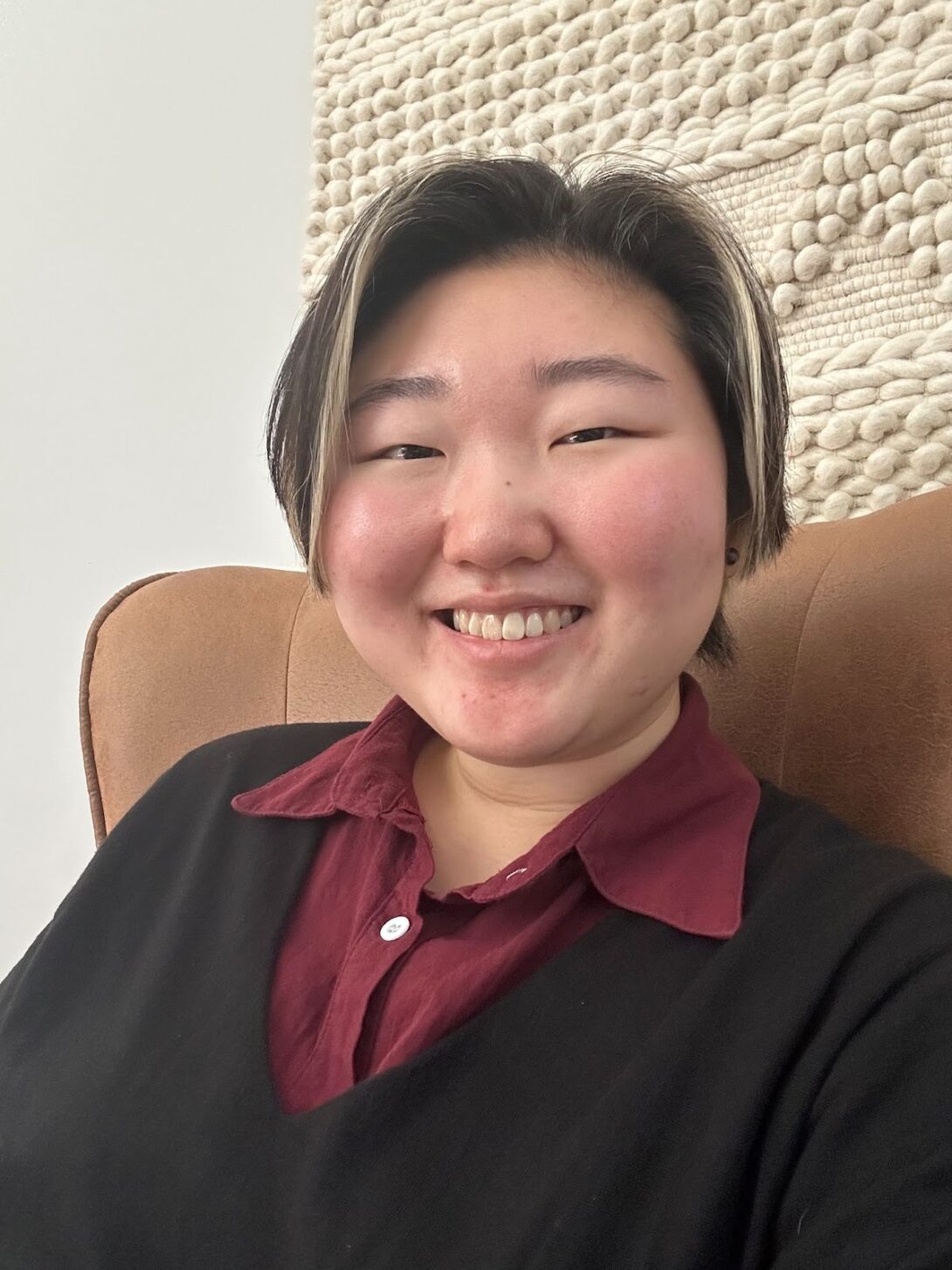 Image Description: a smiling, fair-skinned Korean person with blonde highlights on black, short hair.  They are wearing a maroon collared shirt underneath a black v-neck sweater, with a light brown chair and white knit wall piece in the background.
