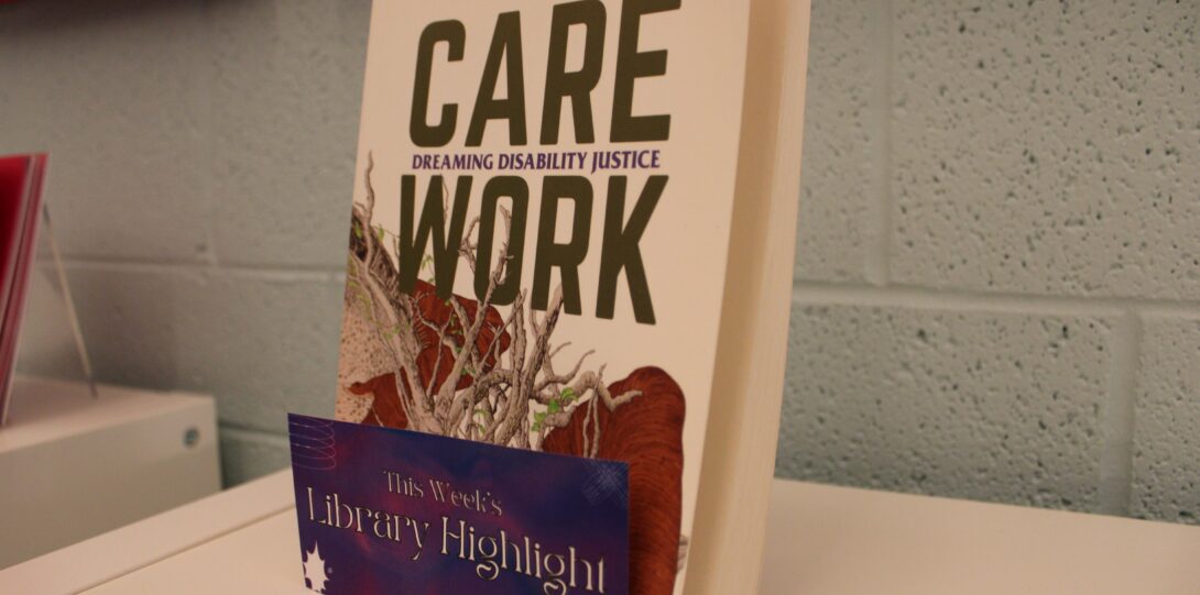 A paperback book displayed upright on top of a white bookshelf in front of a light blue wall. The books is Care Work: Dreaming Disability Justice by Leah Lakshmi Piepzna-Samarasinha. There is a dark purple and pink sign on the display that says 