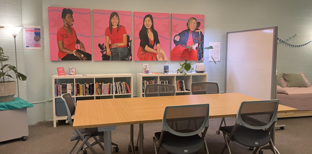 An open room with high ceilings, brown carpet, and light blue cinderblock walls. There are four bright pink Crip Power Paint paintings hung on the wall in the center above short white bookshelves filled with books and plants on top. There is a long light brown table with blue chairs in the middle and a light pink daybed in the right corner behind a large whiteboard divider.