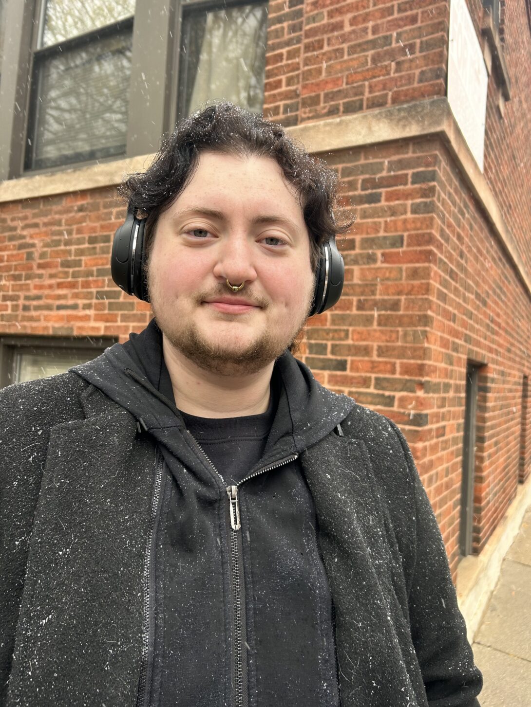 Theo is a white nonbinary person with a beard smiling for the camera. They are standing outside in front of a brick building while it is snowing. They are wearing a black jacket, over the ear headphones, and a large, silver septum ring.